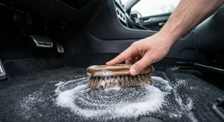 A close-up shot of a hand scrubbing car carpet fabric with a wooden stiff-bristle upholstery detailing brush and white cleaning foam 
